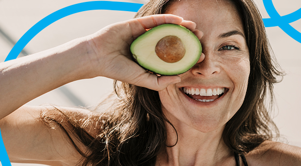 A woman holds half an avocado in front of her eye.