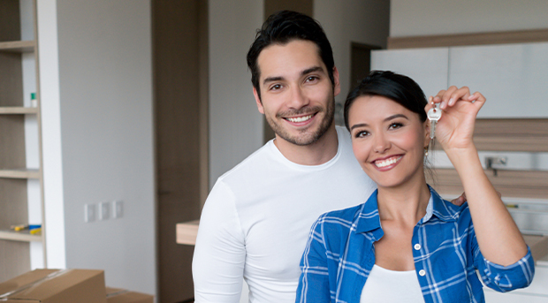 Two first time home owners, a young man in a white shirt and a woman in a blue plaid shirt, smile and hold the keys to their first home, standing in their kitchen with moving boxes behind them