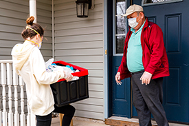 A woman wearing a mask dropping off a box for an older man, also wearing a mask.