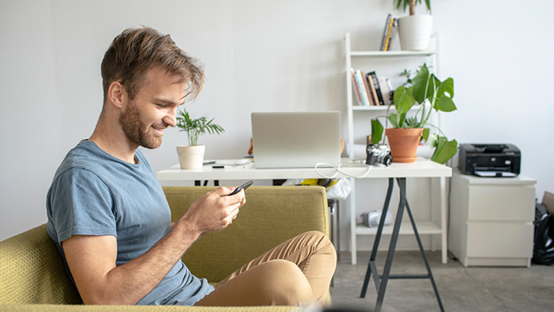 A man sits on his couch smiling and using his personal finance apps on his phone.