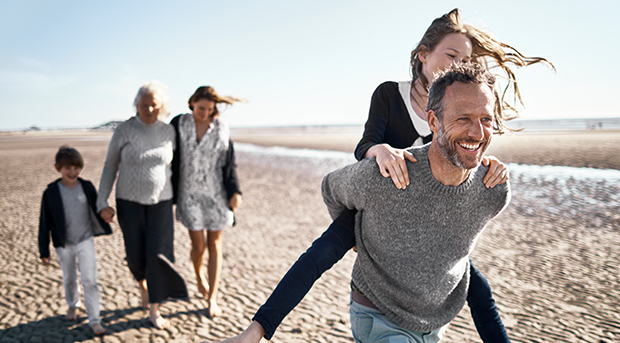 A father carries his daughter on his back at the beach while the rest of his family follow behind them.