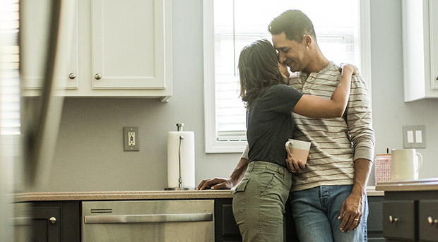 Two first time home owners, a young man in a white shirt and a woman in a blue plaid shirt, smile and hold the keys to their first home, standing in their kitchen with moving boxes behind them