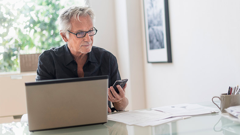 A man sits at his kitchen table using his phone and laptop to bank securely.