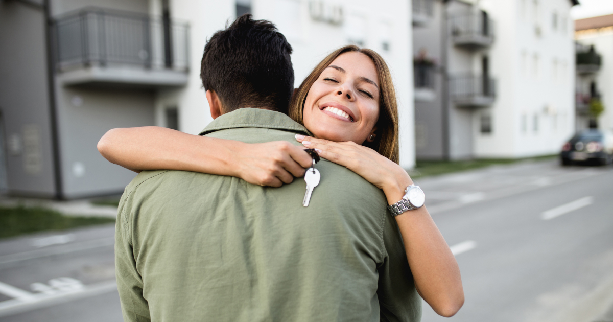 A smiling young woman holding house keys on the street of her new home. She is hugging a man with his back to the camera.