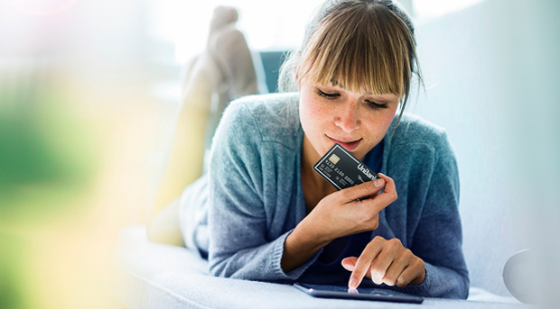 A woman is lying down and looking at a black UniBank Credit card she is holding. Blurred greenery is on her left.