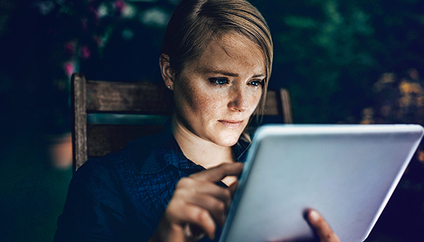 A close up image of a serious-looking woman using a tablet device while sitting in a chair. The background is dark.