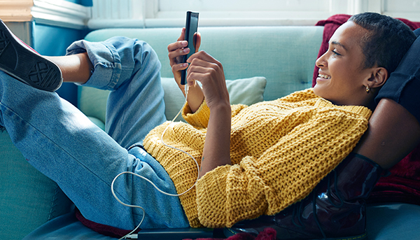 A close up image of a young woman wearing jeans and a yellow jumper. She is smiling and looking at a mobile phone while lying on a couch.