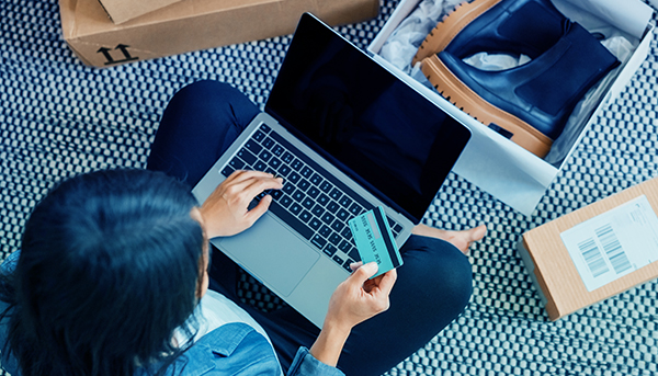 An overhead shot of a person sitting cross-legged on the floor, holding a bank card and using a laptop. An open shoe box with boots inside sits in front.