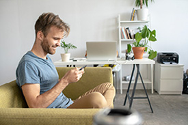 A man sits on his couch smiling and using his personal finance apps on his phone.