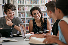 A group of young people sit at a table in the library with their books and laptops, laughing with each other.
