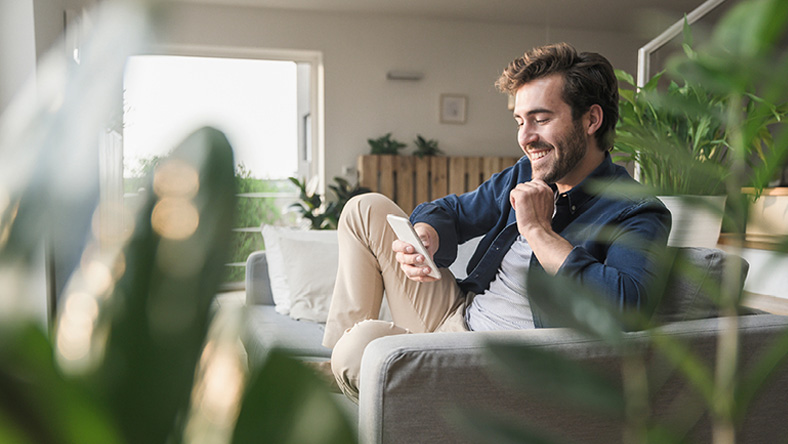 A young man sits on his living room couch surrounded by houseplants, smiling as he reads information on his phone.