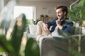 A young man sits on his living room couch surrounded by houseplants, smiling as he reads information on his phone.