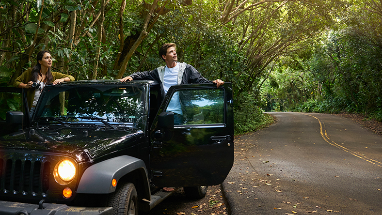 A young woman and young man lean out of the front doors of their new car in a country road.