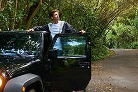 A young woman and young man lean out of the front doors of their new car in a country road.