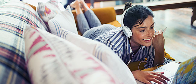 Young woman lying on the couch wearing headphones, using her laptop to create a budget.