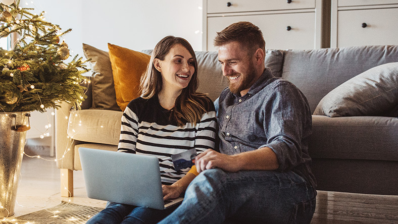 A man and woman sit on their livnig room floor, using a laptop and smiling. A Christmas tree is to their left.