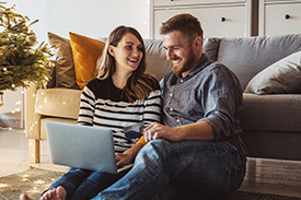 A man and woman sit on their livnig room floor, using a laptop and smiling. A Christmas tree is to their left.