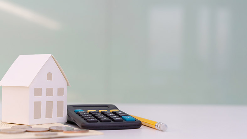 Image of a white desk surface with a small model house, a calculator, a yellow pencil and some coins in focus. The background is plain.