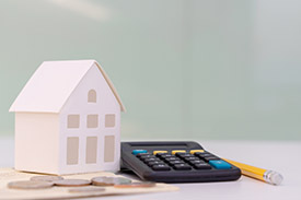 Image of a white desk surface with a small model house, a calculator, a yellow pencil and some coins in focus. The background is plain.