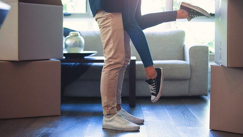 A man lifts his partner's feet in the air as they celebrate in their first home.