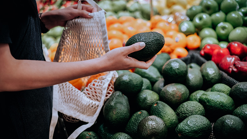 A hand grasps an avocado to place it in a shopping bag in a grocery store
