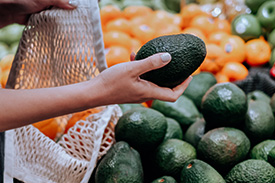 A hand grasps an avocado to place it in a shopping bag in a grocery store