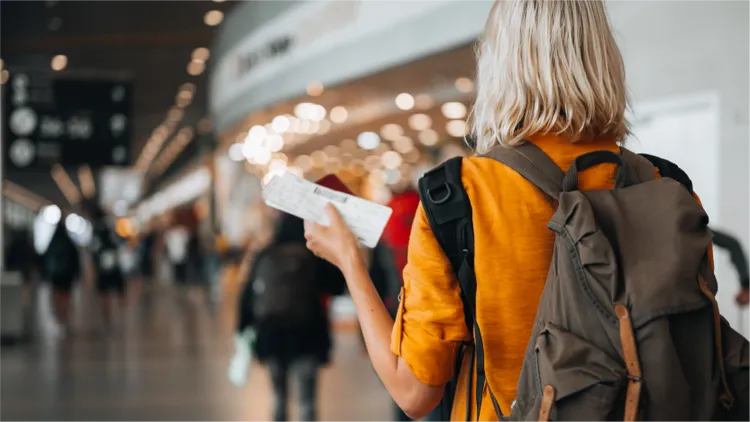 A traveler with a backpack holds a boarding pass while walking through a modern airport terminal.