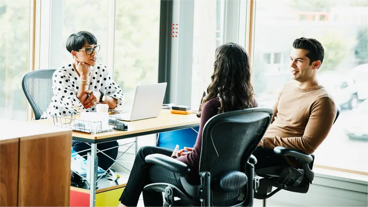 Three colleagues sit together in a modern office space, engaged in a discussion around a desk.