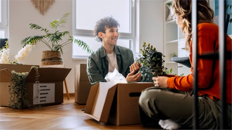 A young couple sits on the floor of a bright apartment surrounded by open cardboard boxes.
