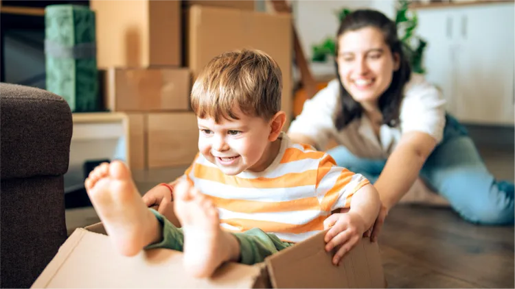 A young child sits inside a cardboard box, being playfully pulled by an adult in a home setting.