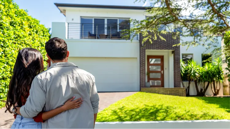 A young couple stands arm in arm, looking toward a modern two-story house.