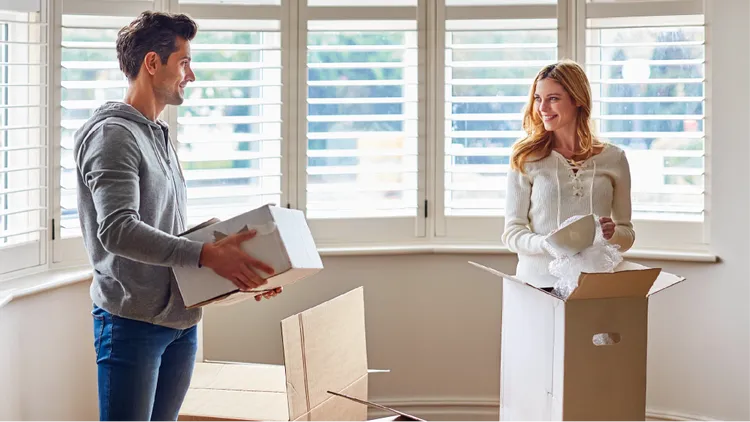 A young couple stands in a bright living room surrounded by cardboard boxes.