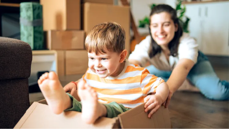 A young child sits inside a cardboard box, being playfully pushed across the floor.