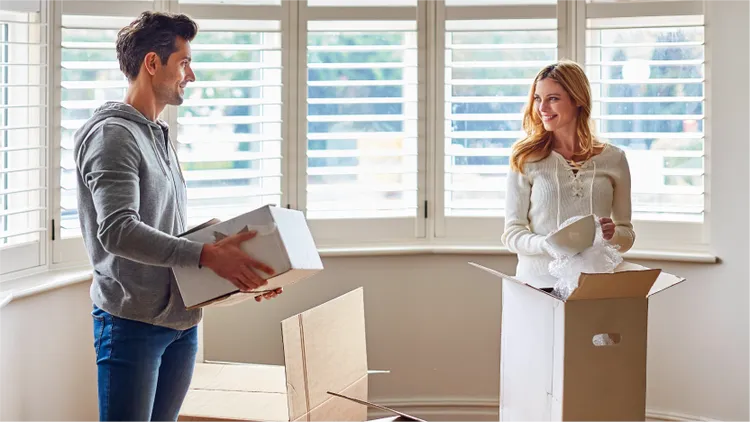 A young couple stands in a bright room filled with moving boxes.