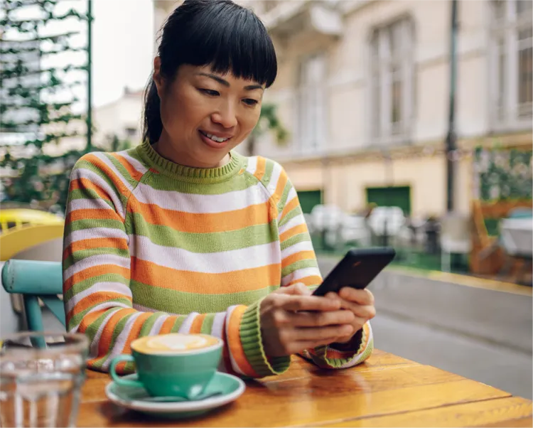 A woman sits at an outdoor cafe table using a smartphone.