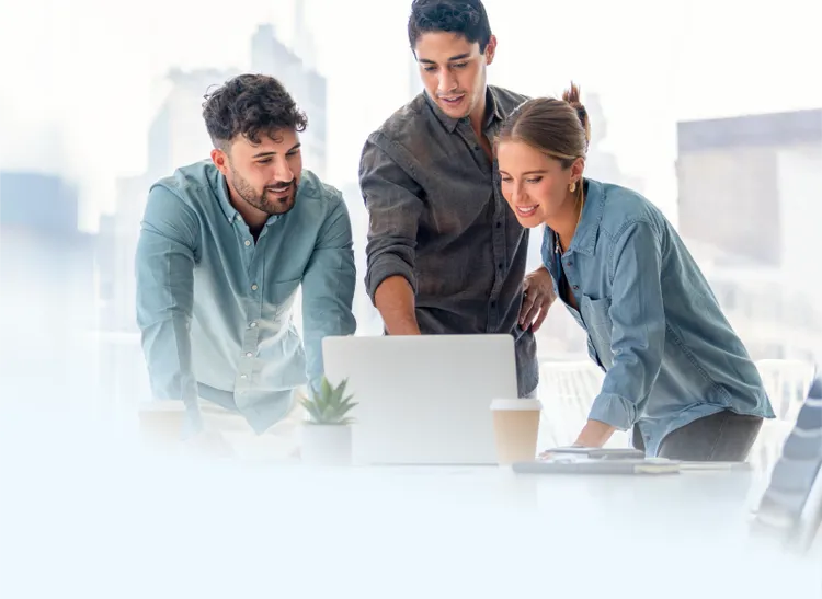 Three colleagues gather around a laptop on a desk in a bright, modern office.