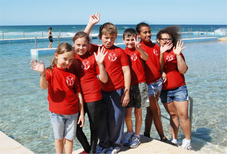 A group of children in matching red t-shirts pose and wave beside a seaside swimming pool.