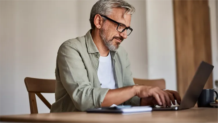 A mature man sits at a wooden table typing on a laptop computer.